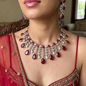 May include: A jewelry set consisting of a necklace and matching drop earrings. The necklace features multiple pear-shaped red gemstones surrounded by small white stones in a metallic setting. The earrings mirror this design with a single red stone.