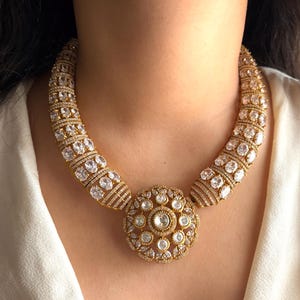 May include: A gold-toned necklace with a large, circular pendant and a curved collar. The necklace is encrusted with numerous clear, sparkling stones. The pendant features a central stone surrounded by smaller stones, creating a radiant effect. The collar has a repeating pattern of stones.