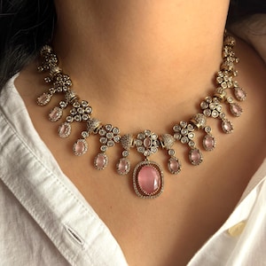 Puede incluir: Un collar dorado con detalles florales y en forma de lágrima, cada uno adornado con pequeñas piedras transparentes y gemas ovaladas de color rosa claro. El collar se lleva sobre una camisa blanca.