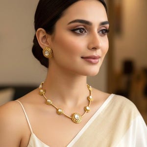 May include: A gold-toned necklace and matching earrings set. The necklace features a series of circular pendants with a central stone, connected by delicate links. The earrings mirror the design. The jewelry is worn with a white top.