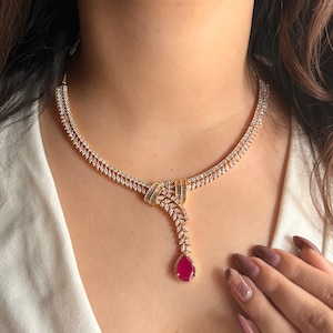 May include: A gold necklace with a teardrop-shaped pendant set with a large red gemstone. The necklace is adorned with small, clear cubic zirconia stones.