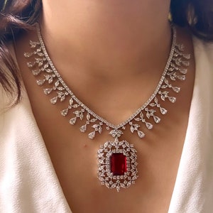 May include: A silver necklace with a large red gemstone pendant surrounded by smaller clear gemstones. The necklace has a delicate, intricate design.