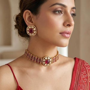 May include: A gold-toned jewelry set featuring a choker necklace and matching stud earrings. The set includes a large, square, ruby-colored stone surrounded by smaller white stones. The necklace and earrings have a detailed, ornate design.