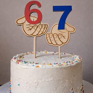 Puede incluir: Un pastel redondo con glaseado blanco y chispas de colores, decorado con dos adornos de madera. Cada adorno muestra una mano sosteniendo un número, con un 6 rojo a la izquierda y un 7 azul a la derecha, ambos montados sobre palitos.