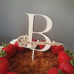 May include: A wooden cake topper with the letter "B" and the name "Benjamin" written in cursive. The topper is placed on a cake decorated with strawberries.