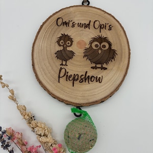 May include: A wooden round sign with the text "Omi's und Opi's Piepshow" and two cartoon owls. A green mesh ball filled with birdseed hangs from the sign.