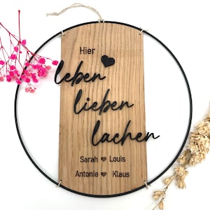 May include: A wooden wall hanging with the words "Hier leben lieben lachen" and names Sarah, Louis, Antonia, and Klaus. The wooden plaque is set within a black metal ring, with a natural twine hanger.
