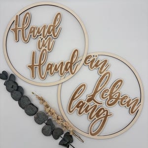 Könnte beinhalten: Zwei kreisförmige Holzdekorationen mit den Worten "Hand in Hand" und "ein Leben lang" in Schreibschrift. Die Dekorationen sind hellbraun und vor einem weißen Hintergrund platziert. Eukalyptus und Trockenblumen sind im Bild.