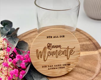 May include: A clear glass tumbler with a wooden lid. The lid is engraved with the words "Für all die kleinen Momente, die das ganz große ausmachen." The tumbler sits on a wooden tray, with pink and dried flowers in the foreground.