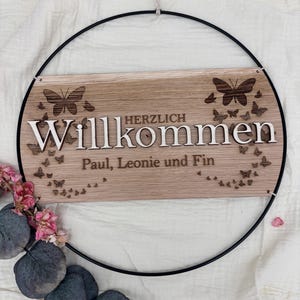 Op de afbeelding: Een rechthoekig houten bord opgehangen in een zwarte metalen ring, voorzien van de gegraveerde tekst "HERZLICH Willkommen" en "Paul, Leonie und Fin" met lasergesneden vlindermotieven rondom de letters en roze bloemen.