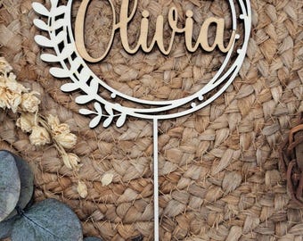 May include: A wooden cake topper with the name "Olivia" in a cursive font, surrounded by a decorative wreath. The topper includes the text "Erstkommunion 29.05.2025" and a fish symbol. The background is a woven texture.