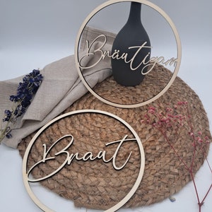 Peut inclure: Deux pancartes rondes en bois avec les mots "Braut" et "Bräutigam" écrits en cursive. Les pancartes sont sur un tapis de paille tressé.