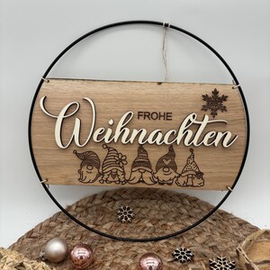 Puede incluir: Un letrero de madera con el texto "Frohe Weihnachten" en una fuente decorativa, rodeado por un anillo de metal negro. El letrero presenta cinco gnomos con sombreros rojos y barbas blancas.