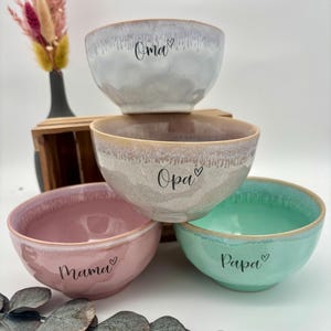 Pode incluir: Uma pilha de quatro tigelas de cerâmica em cores pastel: cinza claro, bege, rosa e verde menta. Cada tigela tem uma inscrição única: "Oma", "Opa", "Mama" e "Papa", com um símbolo de coração. As tigelas têm um acabamento texturizado e esmaltado.