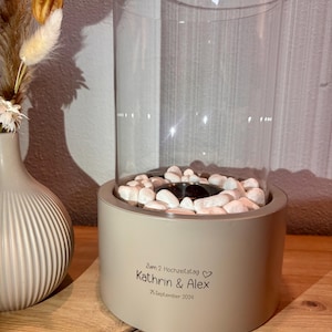 Op de afbeelding: Een cilindervormige beige betonnen tafelhaard met een heldere glazen cilinder en witte decoratieve stenen. De voet is voorzien van de zwarte tekst: Zum 2. Hochzeitstag, Kathrin & Alex, 25. September 2024, inclusief een klein hartje.