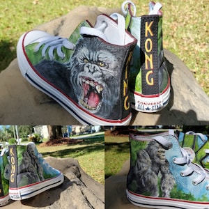 May include: Custom-painted Converse All Star high-top sneakers featuring a detailed illustration of King Kong. The design includes the gorilla's face on the sides and the word "KONG" on the back. The shoes have white laces and soles with a red stripe.