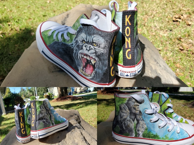 May include: Custom-painted Converse All Star high-top sneakers featuring a detailed illustration of King Kong. The design includes the gorilla's face on the sides and the word "KONG" on the back. The shoes have white laces and soles with a red stripe.