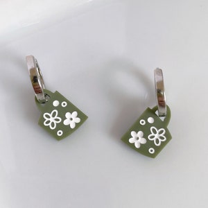 May include: A pair of green earrings with white floral designs. The earrings are shaped like a mug and have silver hoops.