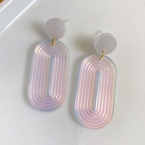 May include: A pair of iridescent oval earrings with a layered design. The earrings are made of a translucent material with a subtle pink hue. The earrings are attached to a small, round, white post.