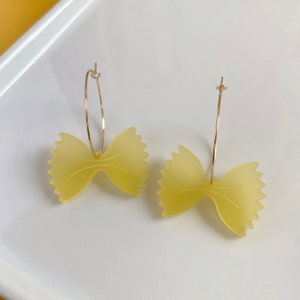 以下が含まれることがあります： 黄色い蝶ネクタイパスタの形をしたユニークなイヤリングのペア。金色のフープから吊り下げられています。イヤリングは白い表面に表示され、遊び心のあるデザインと色を示しています。