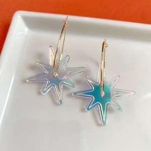 May include: A pair of gold hoop earrings featuring iridescent starburst pendants. The starbursts have a shimmering, multi-coloured effect, reflecting hues of blue, green, and purple. The earrings are displayed on a white surface.