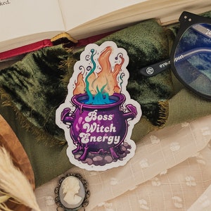 May include: A sticker of a purple cauldron with flames and green smoke. The cauldron has the text "Boss Witch Energy" on it.