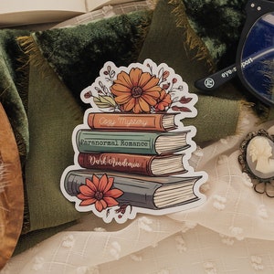 May include: A sticker of a stack of books with floral accents. The books are labeled "Cozy Mystery", "Paranormal Romance", and "Dark Academia".