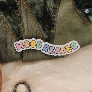 May include: A still-life image featuring a book, a green velvet book rest, and a sticker that reads "MOOD READER" in colorful letters. Also present are a cameo brooch, glasses, and a wooden bowl with dried flowers.