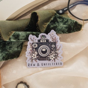 May include: A sticker with a vintage camera surrounded by flowers and the text "RAW & UNFILTERED".