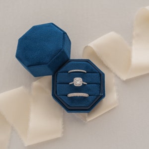 May include: A blue velvet ring box with three rings inside. The box is open and the rings are visible. The box is sitting on a white surface with a white ribbon.