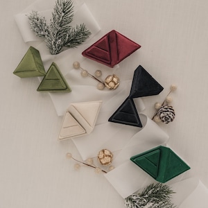 May include: Five velvet ring boxes in different colors, including green, red, cream, black, and dark green, are arranged on a white surface with white ribbon and sprigs of greenery.