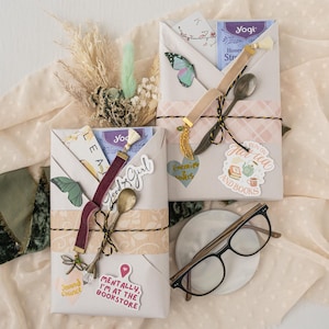 May include: Gift boxes wrapped in cream paper with decorative elements. Each box is adorned with stickers, ribbons, and small spoons. One box has a butterfly sticker. A pair of eyeglasses and a small ceramic dish are also present.