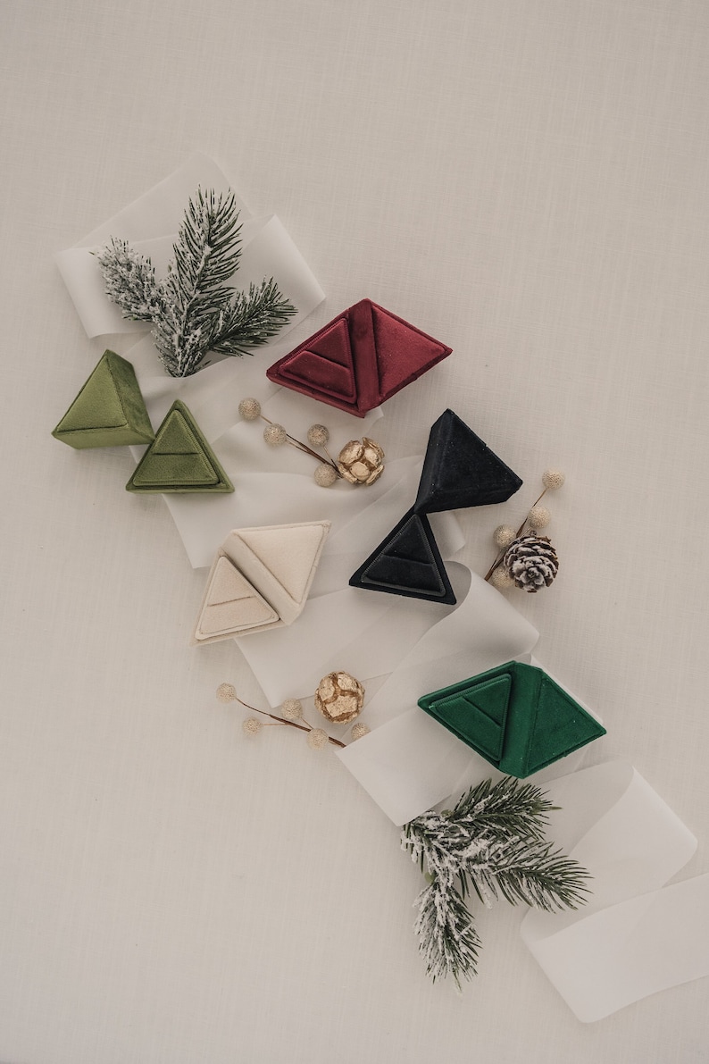 May include: Five velvet ring boxes in different colours, including green, red, cream, black, and dark green, are arranged on a white surface with white ribbon and sprigs of greenery.