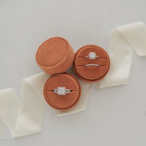May include: Three round velvet ring boxes in a burnt orange color. Two boxes contain engagement rings with a diamond center stone. The third box contains a wedding band.