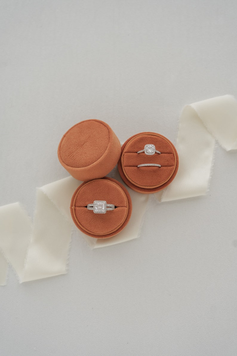 May include: Three round velvet ring boxes in a burnt orange color. Two boxes contain engagement rings with a diamond center stone. The third box contains a wedding band.