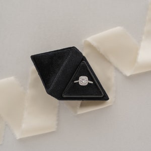 May include: A black velvet ring box shaped like a pyramid, containing a silver ring with a large square-shaped diamond.