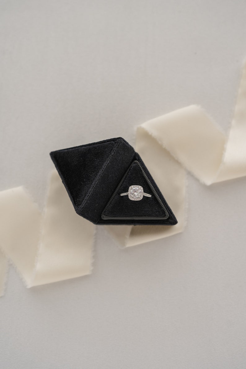 May include: A black velvet ring box shaped like a pyramid, containing a silver ring with a large square-shaped diamond.