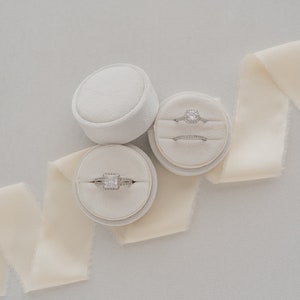 May include: Two white ring boxes with velvet interiors, each containing a silver ring with a clear diamond. The boxes are on a white surface with a cream-colored ribbon.