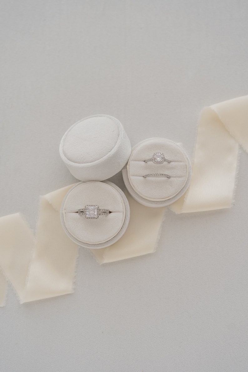 May include: Two white ring boxes with velvet interiors, each containing a silver ring with a clear diamond. The boxes are on a white surface with a cream-colored ribbon.