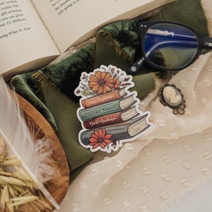 May include: A sticker of a stack of books with floral accents. The books are labeled "Cozy Mystery", "Paranormal Romance", and "Dark Academia".