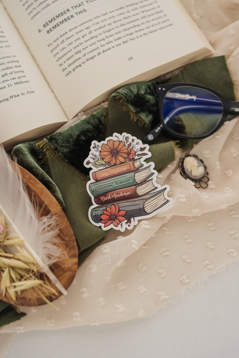 May include: A sticker of a stack of books with floral accents. The books are labeled "Cozy Mystery", "Paranormal Romance", and "Dark Academia".