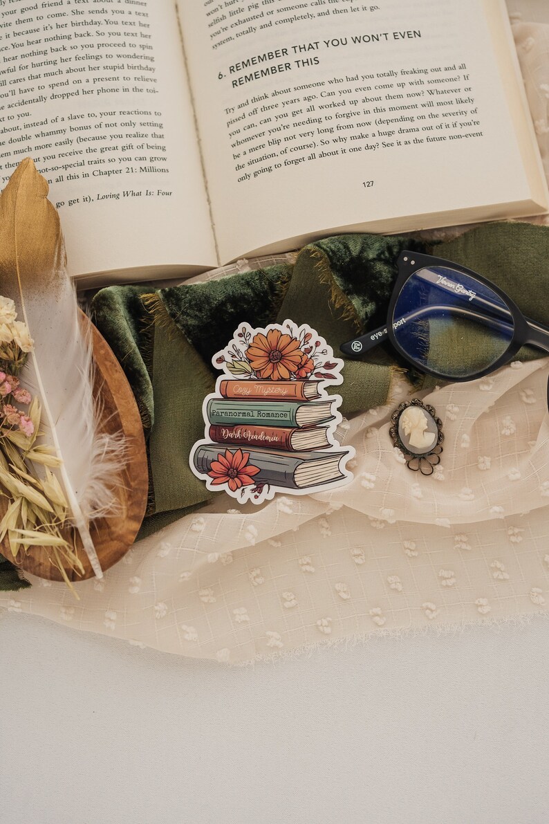 May include: A sticker of a stack of books with floral accents. The books are labeled "Cozy Mystery", "Paranormal Romance", and "Dark Academia".