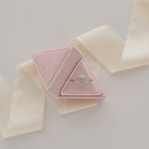 May include: A pink velvet ring box shaped like a pyramid, containing a silver ring with a square-shaped diamond.