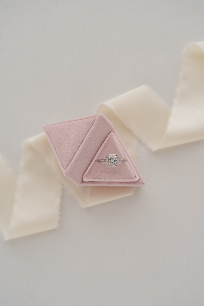 May include: A pink velvet ring box shaped like a pyramid, containing a silver ring with a square-shaped diamond.