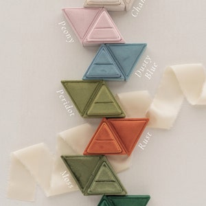 May include: Nine triangular ring boxes in a variety of colours, including almond, champagne, peony, dusty blue, peridot, rust, moss, pine, and jet. Each box is made of velvet and has a ring-shaped indentation in the centre.