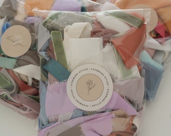 May include: Several clear plastic bags filled with colorful fabric scraps. The fabrics are in shades of blue, green, orange, and purple. Each bag has a round sticker with the text "EVERMORE STYLED" and a floral design.