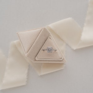 May include: A white velvet ring box shaped like a pyramid, containing a silver ring with a large, clear, square-shaped diamond.