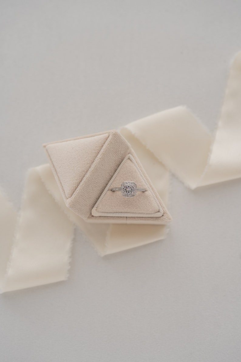 May include: A white velvet ring box shaped like a pyramid, containing a silver ring with a large, clear, square-shaped diamond.
