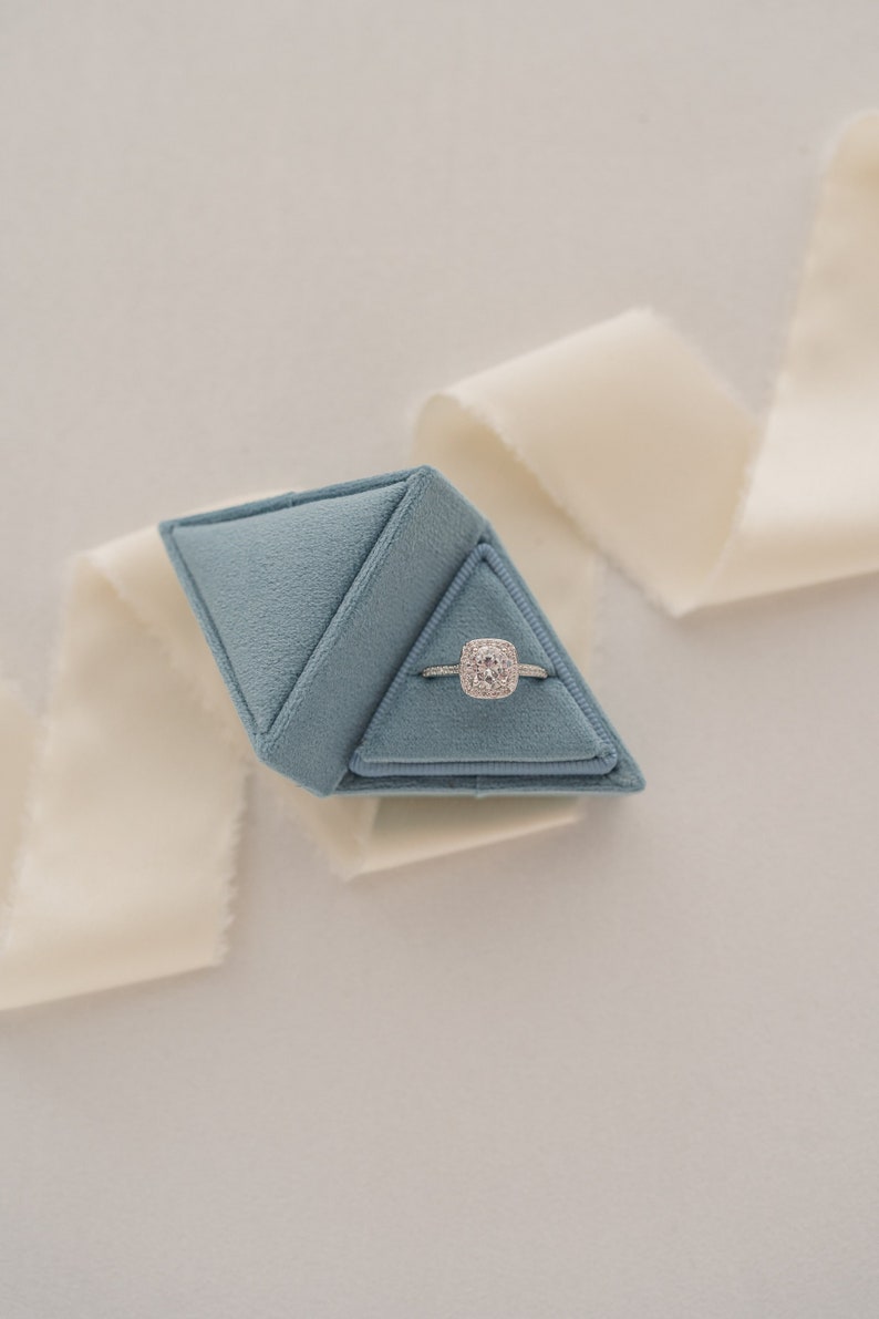 May include: A blue velvet ring box shaped like a pyramid, containing a silver ring with a large, square-shaped diamond.