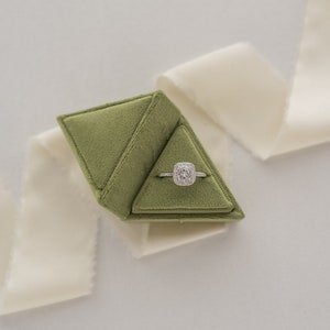 May include: A green velvet ring box with a diamond engagement ring inside. The box is shaped like a pyramid and is sitting on a white surface.